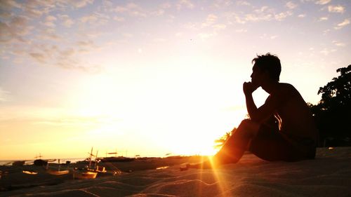Side view of thoughtful man sitting at sandy beach against sky during sunset