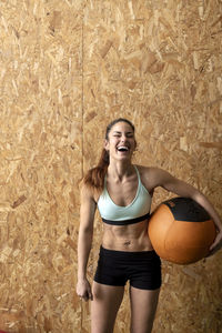 Beautiful smiling young female in sportswear holding heavy leather ball and looking at camera while standing near cork wall at gym