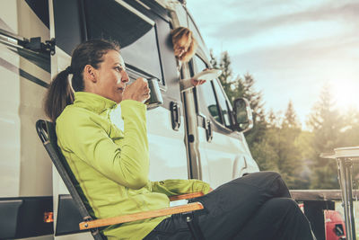 Side view of smiling woman drinking coffee while sitting by motor home