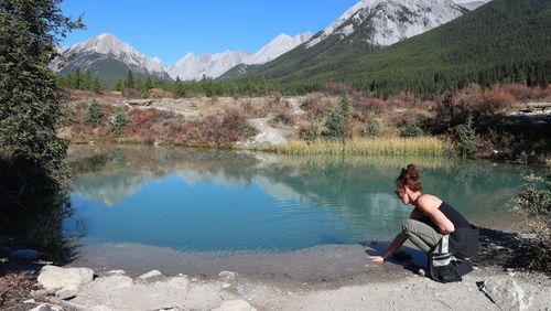 Side view of woman crouching on land by lake against mountains