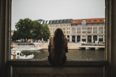 Rear view of woman sitting on window sill against buildings in city