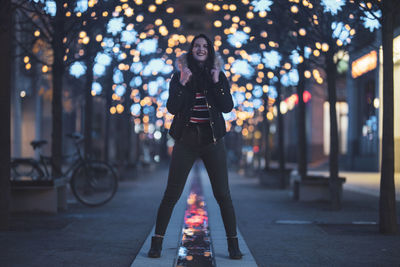 Woman standing on footpath against illuminated lights at night