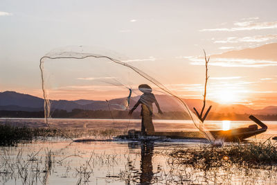 Silhouette fisherman throwing fishing net in lake against sky during sunset