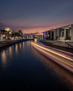 Light trails on river by illuminated buildings against sky at night