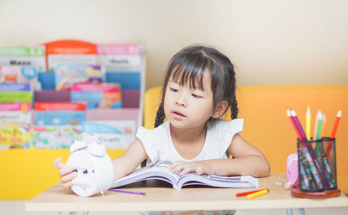 Cute girl holding clock while reading book on table