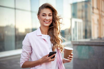 Portrait of smiling woman holding smart phone while standing in city