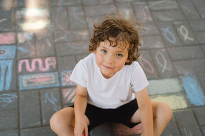 Little preschool boy draws with colorful chalks on the ground.