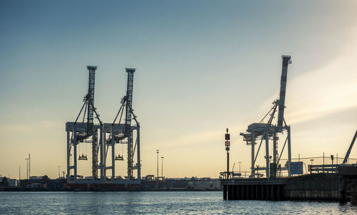 Cranes at harbor against sky during sunset