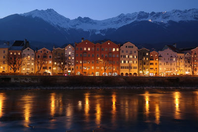 Illuminated buildings by river against mountain during winter in town at dusk