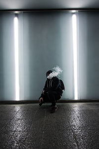 Man exhaling smoke while crouching against wall