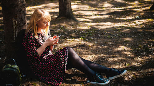 Woman using mobile phone while sitting by tree trunk in forest