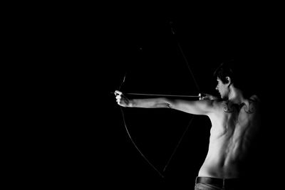 Rear view of naked woman aiming bow against black background