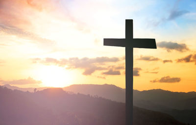 Silhouette cross on mountains against cloudy sky during sunset