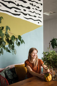 Woman sits on sofa surrounded by plants, looks at camera with smile.