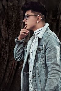 Side view of thoughtful young man wearing denim jacket while standing by tree trunk