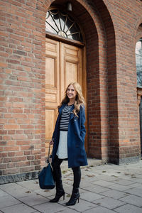 Full length portrait of smiling young woman standing on footpath against building in city