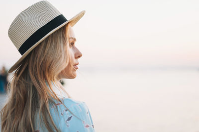 Portrait of beautiful blonde young woman in blue dress and straw hat on pier on sunset