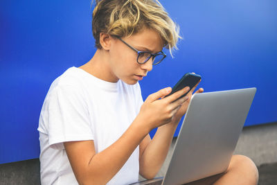 Boy using mobile phone while sitting against wall