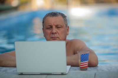 Mature man looking at laptop while swimming in pool