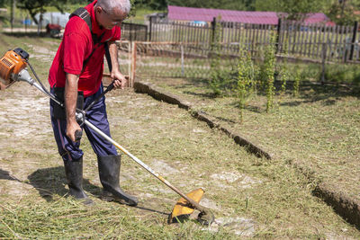 A mature man with a trimmer cuts grass and weeds with a trimmer on a paved path.