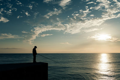 Silhouette man fishing in sea while standing on pier against sky during sunset