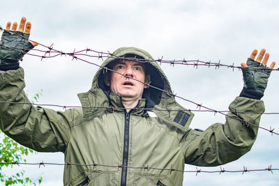 A young guy stands behind a barbed wire fence and holds onto the barbed wire with his hands.