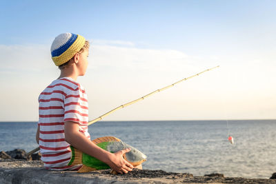 Boy sitting with toy fish on retaining wall while fishing in sea