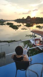 Rear view of woman sitting by swimming pool against lake during sunset