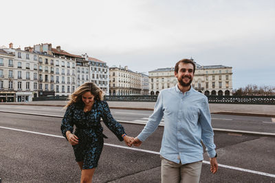 Happy young romantic couple in stylish clothes laughing and holding hands while crossing bridge with historic buildings in background during city tour in bayonne in france