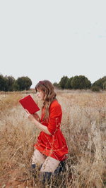 Mid adult woman reading book while kneeling on grassy field against clear sky