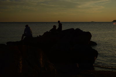 Silhouette people on rock by sea against sky during sunset