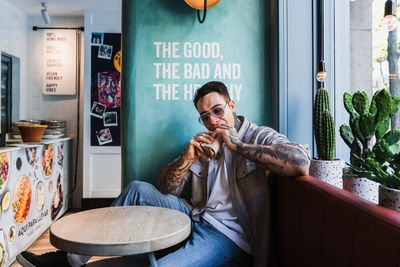 Cool male in sunglasses enjoying delicious takeaway food at table against cacti on windowsill in restaurant