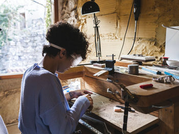 Side view of curly haired ethnic female worker in transparent glasses concentrating at goldsmith workbench working with wire