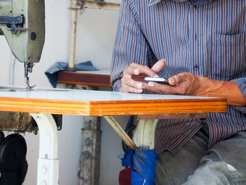 Midsection of man using mobile phone while sitting by sewing machine