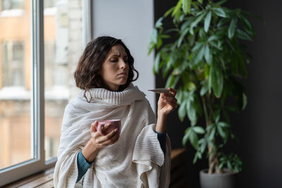 Sick unhealthy young woman with high fever looking at thermometer, checking body temperature