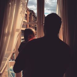 Rear view of man carrying baby by window at home