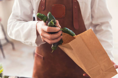 Midsection of salesman putting green chili peppers in paper bag at grocery store