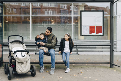 Father feeding baby food to son while sitting with daughter at bus stop in city