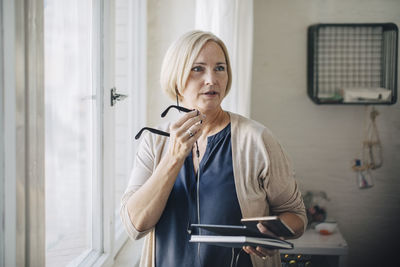 Mature businesswoman talking through earphones while standing by window in office