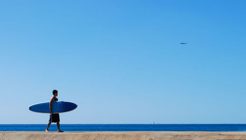 Surfer walking on beach against clear blue sky