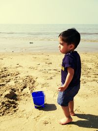Side view of cute boy standing by bucket on sandy beach