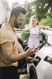 Side view of man using smart phone while charging electric car