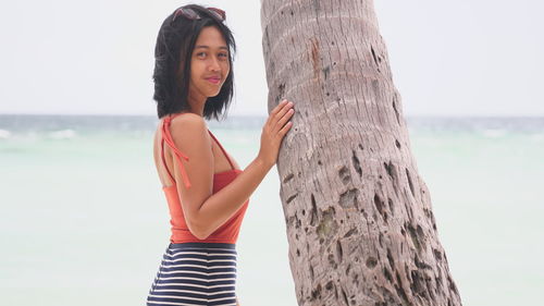 Side view portrait of young woman standing by tree trunk at beach