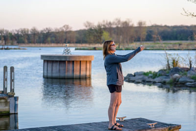 Full length of woman photographing while standing on jetty over lake during sunset