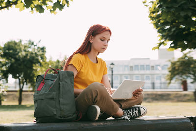 A teenage girl is sitting on a park bench with a laptop and preparing for lessons or exams.