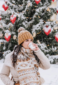 Woman having drink while standing against christmas tree