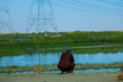 Man sitting on field by lake against sky