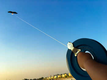 Cropped hand holding spool while flying kite in blue sky