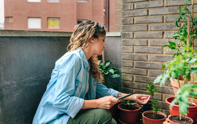 Woman checking plants of urban garden on terrace while holding digital tablet in hand