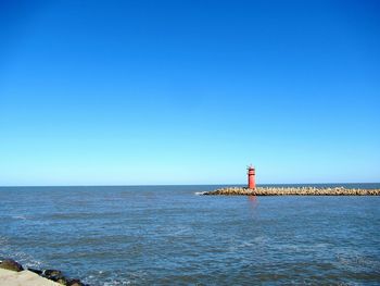 Lighthouse by sea against clear blue sky